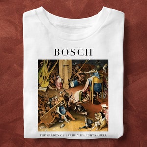 May include: White t-shirt featuring a print of Hieronymus Bosch's painting "The Garden of Earthly Delights - Hell." The word "BOSCH" is printed above the artwork. The shirt is folded and displayed on a reddish-brown surface.