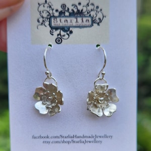 May include: Silver flower dangle earrings with a delicate design. The earrings are on a white card with the text "Starlia Handcrafted Jewellery" and website information.
