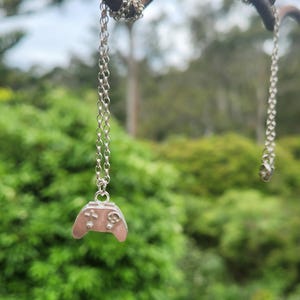 May include: A silver-colored necklace with a small pendant shaped like a video game controller. The controller has a cross-shaped directional pad and buttons. The necklace hangs against a backdrop of green foliage.