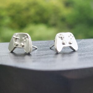 May include: A pair of silver-colored stud earrings shaped like video game controllers. Each controller has a cross-shaped directional pad, four buttons, and two control sticks. The earrings are displayed on a dark surface against a blurred green background.