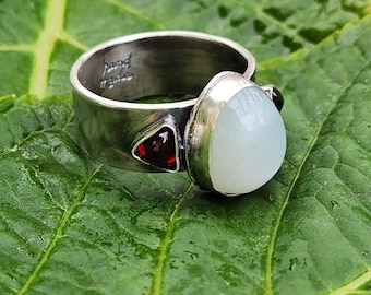 May include: A silver ring with a white oval gemstone and two red triangular gemstones. The ring is engraved with the words "hand made".
