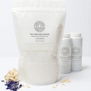 May include: A white bag of talc-free body powder labeled "Lavender" with the text "Shady Acre Homestead" and "Handcrafted in Upstate New York." Two white bottles are behind the bag. Dried lavender and flowers are scattered around the bag.