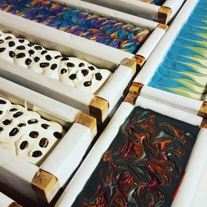 May include: Five bars of handmade soap in wooden molds. The soaps are in various colors and patterns, including white with coffee beans, blue and green swirls, and black with red and orange swirls.