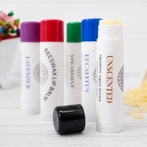 May include: A collection of white lip balm tubes with colorful caps. The tubes are labeled with the scents: Lavender, Beeswax, Spearmint, Eucalyptus, and Unscented. The Unscented tube is open, revealing the balm. A black cap sits in front.