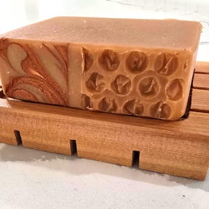 K&ouml;nnte beinhalten: Ein rechteckiger Seifenblock in Hellbraun, der auf einer Holzseifenschale liegt. Die Seife hat ein einzigartiges Design mit kupferfarbenen Wirbeln auf einer Seite und einem Wabenmuster auf der anderen Seite. Die Holzschale hat eine nat&uuml;rliche Oberfl&auml;che.