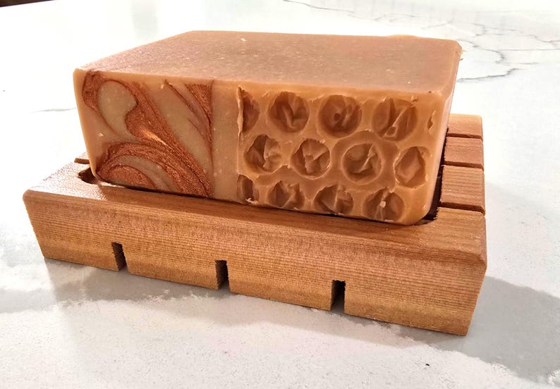 K&ouml;nnte beinhalten: Ein rechteckiger Seifenblock in Hellbraun, der auf einer Holzseifenschale liegt. Die Seife hat ein einzigartiges Design mit kupferfarbenen Wirbeln auf einer Seite und einem Wabenmuster auf der anderen Seite. Die Holzschale hat eine nat&uuml;rliche Oberfl&auml;che.