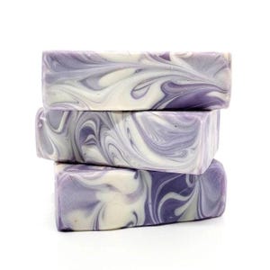 May include: Three stacked bars of handmade soap. Each bar features a swirling pattern of lavender and white, creating a marbled effect. The soap is rectangular and appears to be unscented, with a smooth texture.