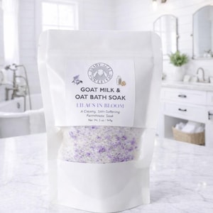 May include: A white pouch of Goat Milk & Oat Bath Soak, labeled "Lilacs in Bloom." The bag contains a mixture of white and purple granules. The net weight is 5 oz.