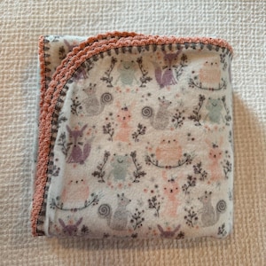 May include: Folded white fleece baby blanket with a repeating pattern of pastel-colored woodland creatures and floral accents. The blanket is trimmed with a crocheted coral edge. A cozy and decorative item for a nursery.