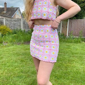 Könnte beinhalten: Zweiteiliges Häkelset in Rosa mit Gänseblümchenmuster. Das Set besteht aus einem Crop-Top und einem Minirock. Die Gänseblümchen haben gelbe Zentren, weiße Blütenblätter und hellblaue Akzente. Das Outfit ist handgefertigt.