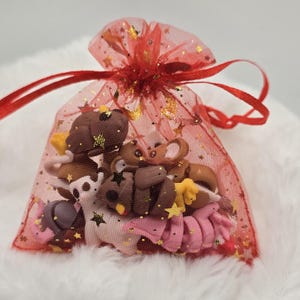 May include: A red mesh gift bag filled with small, colorful clay figurines. The bag is tied with a red ribbon and adorned with gold stars. The figurines include various shapes and colors, such as brown, pink, and yellow, suggesting a craft or handmade theme.
