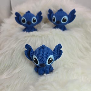 Peut inclure: Trois figurines bleues de Stitch, avec de grandes oreilles et des yeux noirs et blancs, sont pr&eacute;sent&eacute;es sur une surface blanche et duveteuse. Les figurines sont faites d'un mat&eacute;riau tricot&eacute; et sont en position assise.