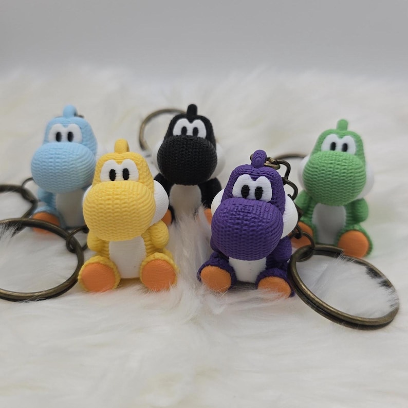 May include: Five colorful Yoshi keychains in blue, yellow, black, purple, and green. Each keychain features a small, detailed Yoshi figure with a round head, large eyes, and a saddle. The keychains are attached to metal rings.