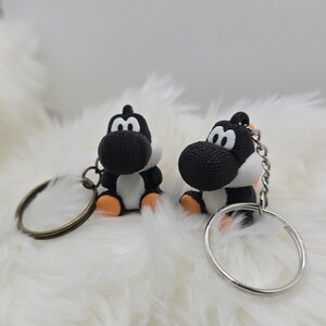 May include: Two black and white Yoshi keychains with orange feet and a silver keyring. The keychains are sitting on a fluffy white surface. The keychains are a cute and fun accessory.