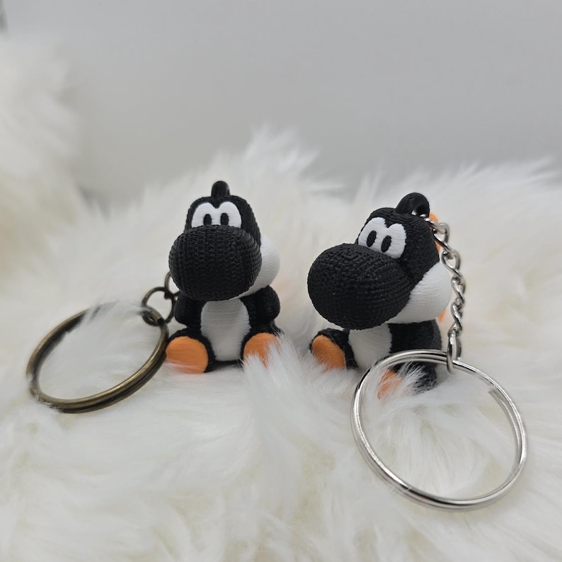 May include: Two black and white Yoshi keychains with orange feet and a silver keyring. The keychains are sitting on a fluffy white surface. The keychains are a cute and fun accessory.