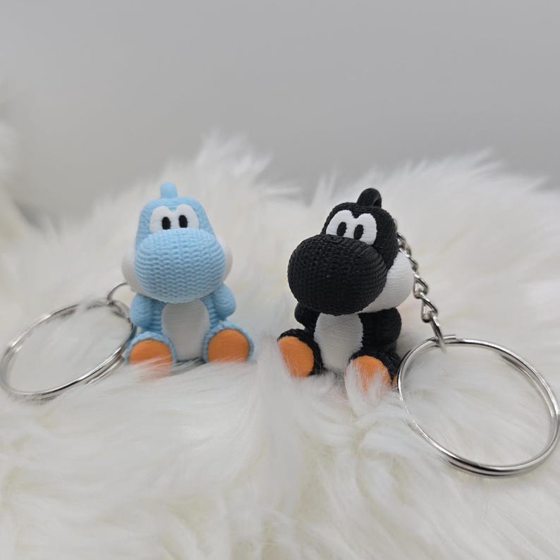 May include: Two character keychains. One is light blue, the other is black and white. Both have orange feet and are attached to silver keyrings. The keychains are sitting on a white, fluffy surface.