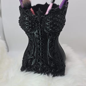 May include: A black corset-shaped organizer featuring intricate embossed scrollwork, a central lace-up detail, and textured bust cups. The container holds four makeup brushes with pink and purple handles, providing a functional storage solution.