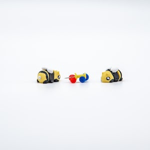 May include: Two yellow and black bee-shaped erasers with white wings are on a white surface. A yellow push pin with red and blue accents is between the erasers. The erasers and push pin are small and appear to be novelty items.