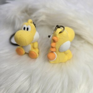 May include: Two yellow Yoshi keychains with white and orange accents. The keychains are made of a textured material and feature a loop for attaching to keys or a bag. One keychain is facing forward, and the other is facing sideways.
