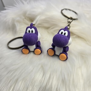 May include: Two purple Yoshi keychains with white bellies, eyes, and orange feet. The keychains are attached to metal rings. The background is a fluffy white surface.