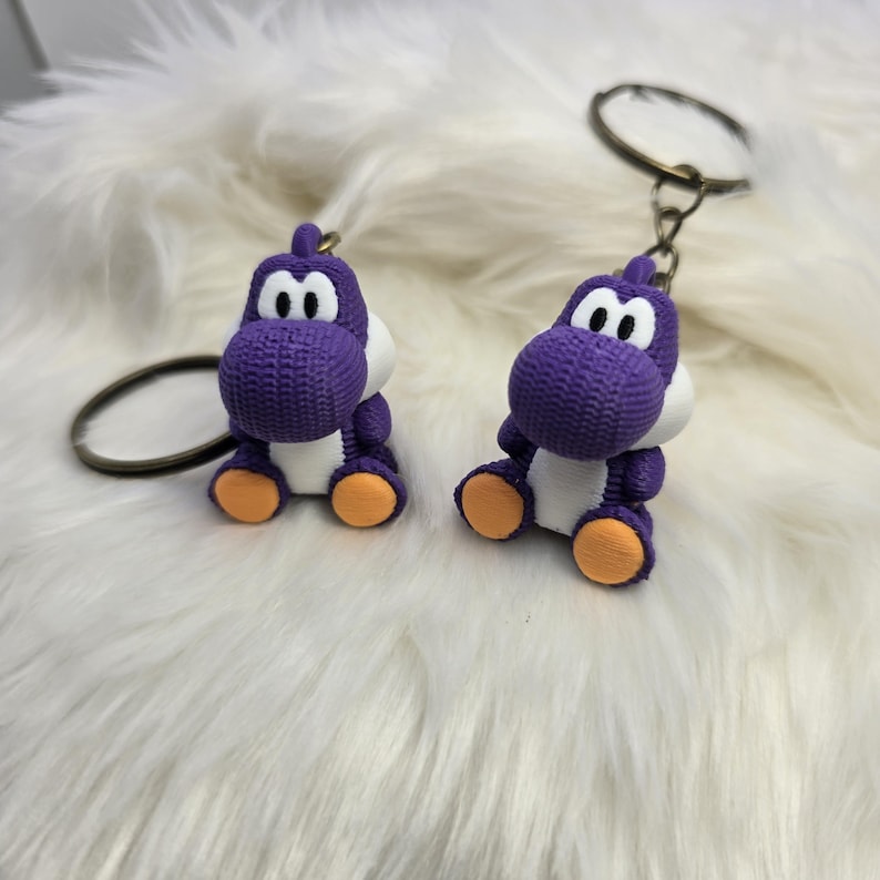 May include: Two purple Yoshi keychains with white bellies, eyes, and orange feet. The keychains are attached to metal rings. The background is a fluffy white surface.