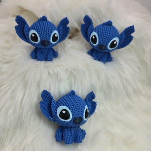 Peut inclure: Trois figurines bleues de Stitch, avec de grandes oreilles et de grands yeux, sont dispos&eacute;es sur une surface blanche et duveteuse. Les figurines sont faites d'un mat&eacute;riau textur&eacute; et ont chacune un petit nez fonc&eacute;.