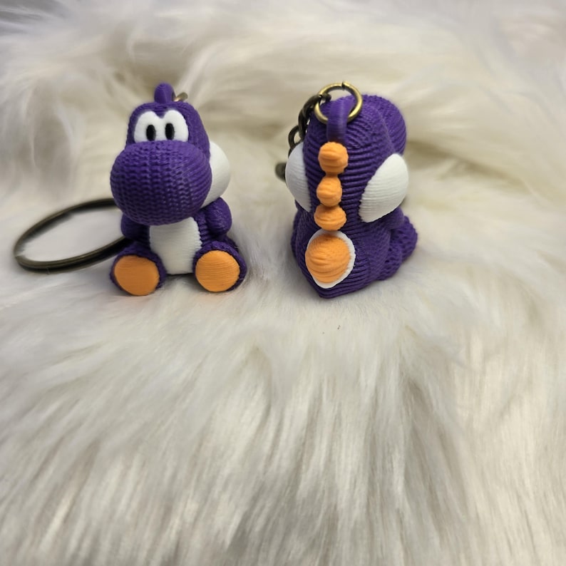 May include: Two purple Yoshi keychains with white bellies, eyes, and accents, and orange feet. The keychains are made of a textured material and have metal key rings.