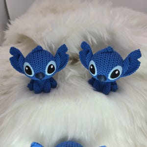 Peut inclure: Deux figurines Stitch bleues tricot&eacute;es, avec de grandes oreilles et des yeux noirs et blancs. Les figurines sont pos&eacute;es sur une surface blanche et duveteuse. Les figurines sont un personnage populaire du film d'animation Lilo & Stitch.