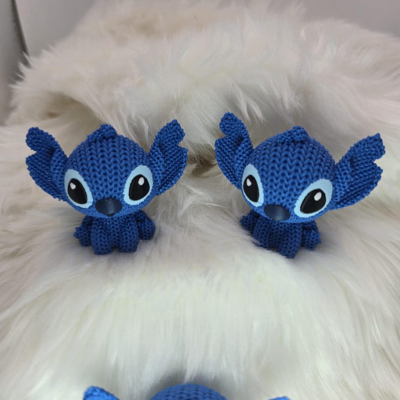 Peut inclure: Deux figurines Stitch bleues tricot&eacute;es, avec de grandes oreilles et des yeux noirs et blancs. Les figurines sont pos&eacute;es sur une surface blanche et duveteuse. Les figurines sont un personnage populaire du film d'animation Lilo & Stitch.