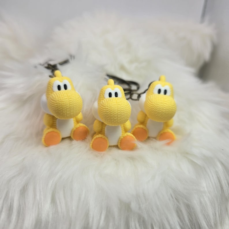 May include: Three yellow Yoshi keychains with white bellies and orange feet are displayed. The keychains have black eyes and are attached to silver key rings. The background is a fluffy white surface.