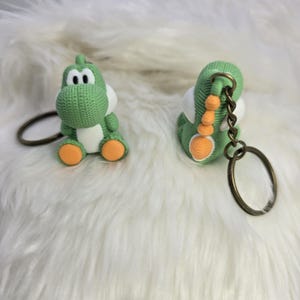 May include: Two green Yoshi keychains with white bellies and orange feet are displayed. The keychains have a knitted texture and are attached to bronze-colored key rings. One keychain is upright, and the other is on its side.