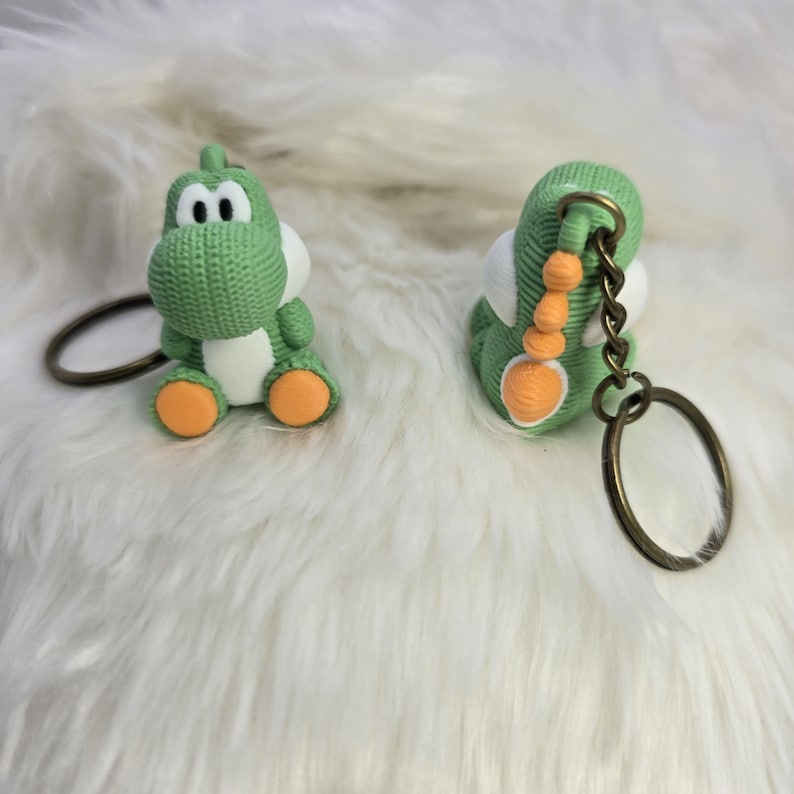 May include: Two green Yoshi keychains with white bellies and orange feet are displayed. The keychains have a knitted texture and are attached to bronze-colored key rings. One keychain is upright, and the other is on its side.
