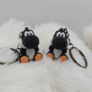 May include: Two black and white Yoshi keychains with orange feet and bronze key rings. The keychains are sitting on a fluffy white surface. The keychains are a popular character from the Super Mario video game series.