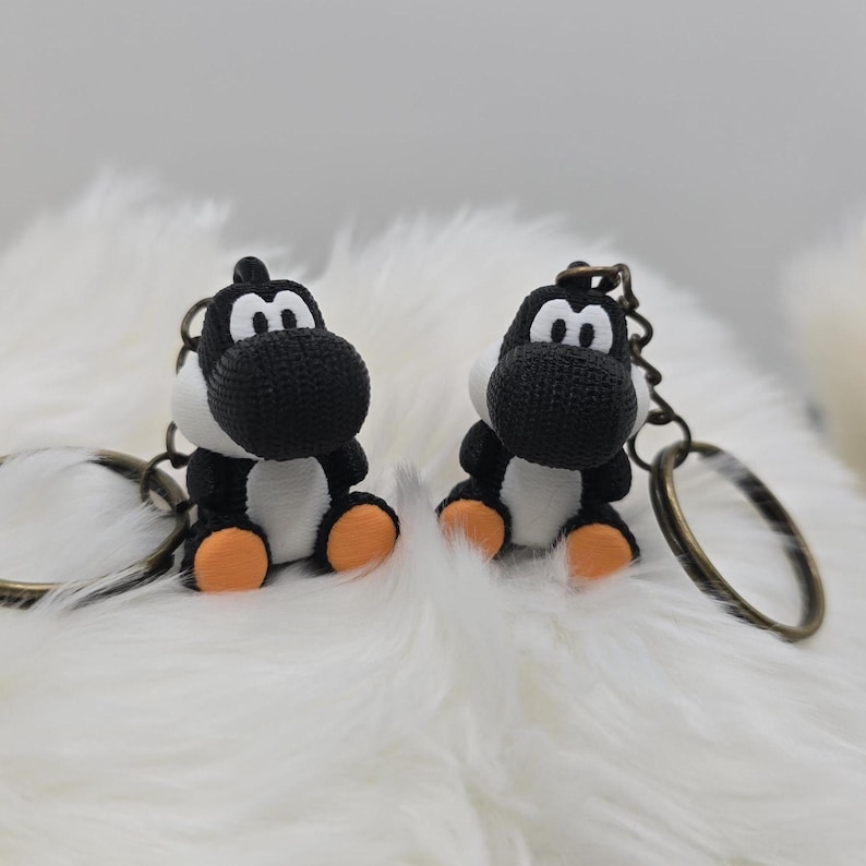 May include: Two black and white Yoshi keychains with orange feet and bronze key rings. The keychains are sitting on a fluffy white surface. The keychains are a popular character from the Super Mario video game series.