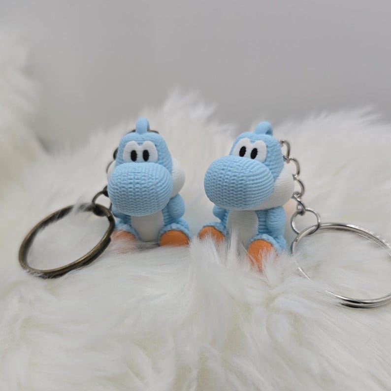 May include: Two light blue Yoshi keychains with white bellies and orange feet are displayed. The keychains have silver metal rings. The background is a soft, white, fluffy surface.