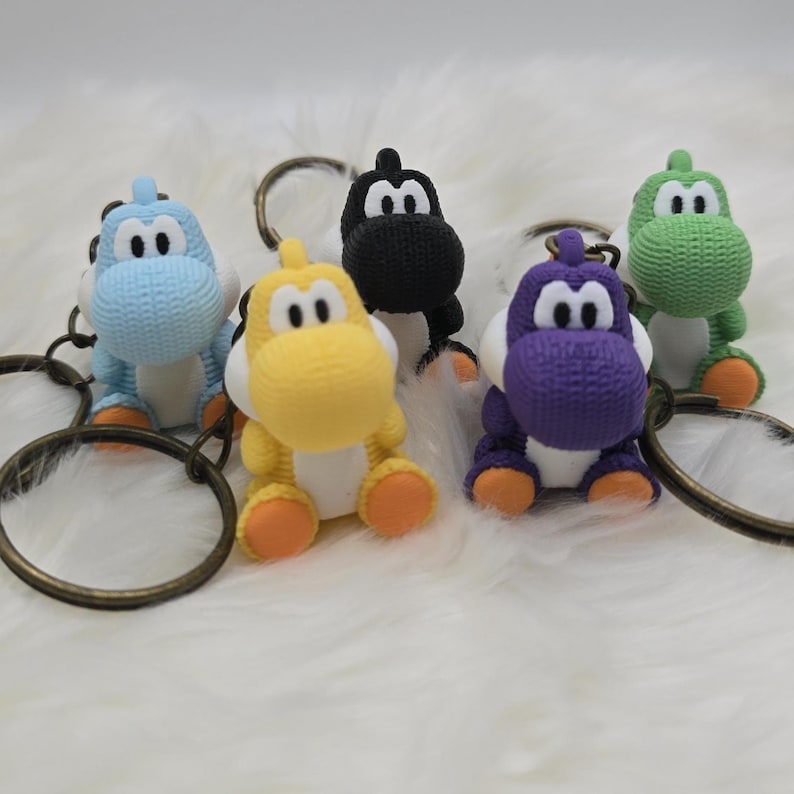 May include: Five colorful Yoshi keychains in blue, yellow, black, purple, and green. Each keychain features a detailed Yoshi figure with a textured appearance, white bellies, and orange feet. The keychains have bronze-colored rings.