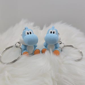 May include: Two blue Yoshi keychains with white bellies and orange feet sit side-by-side. Each keychain has a silver ring. The keychains are on a fluffy white surface.