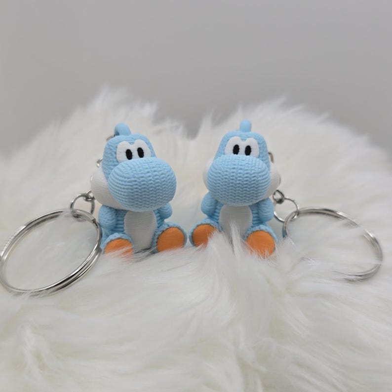 May include: Two blue Yoshi keychains with white bellies and orange feet sit side-by-side. Each keychain has a silver ring. The keychains are on a fluffy white surface.