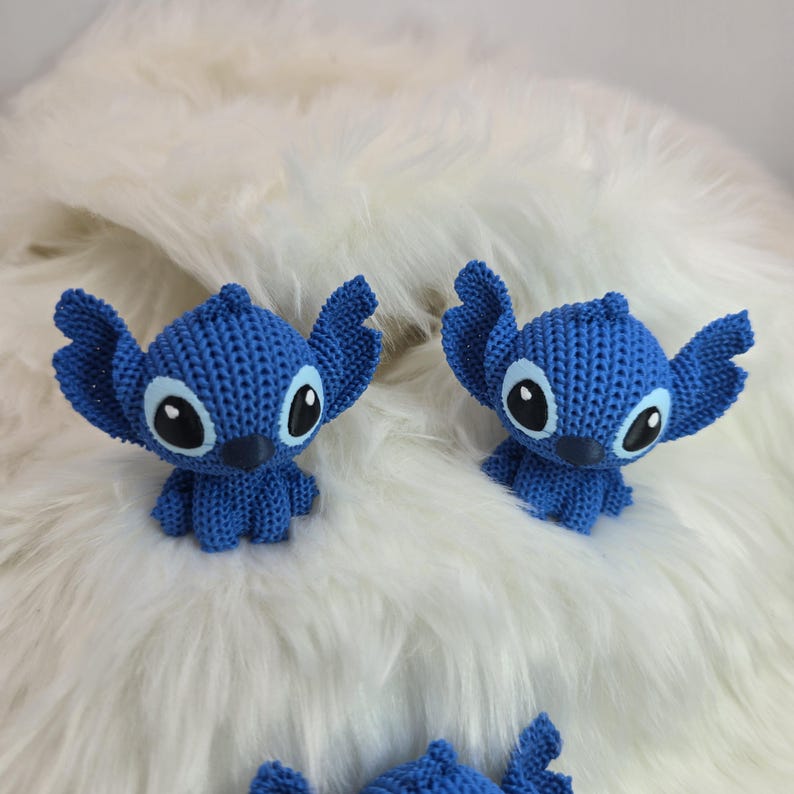 Peut inclure: Plusieurs petites figurines Stitch bleues crochet&eacute;es, avec de grandes oreilles et des yeux noirs et blancs, sont pr&eacute;sent&eacute;es sur une surface blanche et duveteuse. Les figurines sont un personnage populaire d'un film d'animation bien connu.