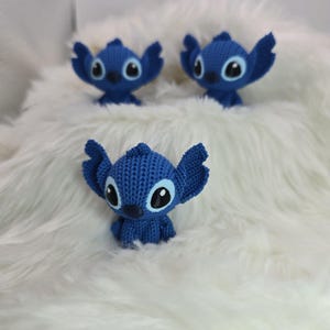 Peut inclure: Trois figurines bleues au crochet repr&eacute;sentant un personnage de dessin anim&eacute; avec de grandes oreilles et des yeux, sur un fond blanc duveteux. Les figurines ont des yeux noirs et blancs et un aspect textur&eacute;.