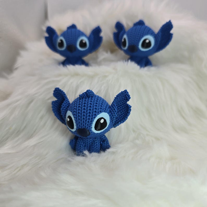 Peut inclure: Trois figurines bleues au crochet repr&eacute;sentant un personnage de dessin anim&eacute; avec de grandes oreilles et des yeux, sur un fond blanc duveteux. Les figurines ont des yeux noirs et blancs et un aspect textur&eacute;.