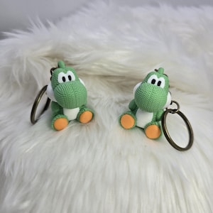 May include: Two green Yoshi keychains with white bellies and orange feet sit on a fluffy white surface. Each keychain has a bronze-colored ring. The Yoshi figures have black eyes and are made of a textured material.