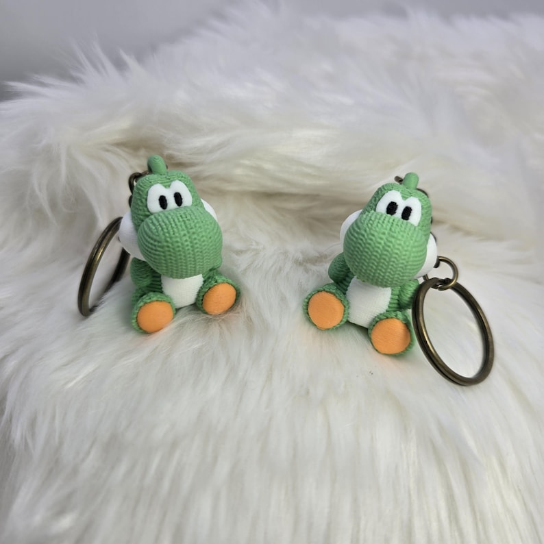 May include: Two green Yoshi keychains with white bellies and orange feet sit on a fluffy white surface. Each keychain has a bronze-colored ring. The Yoshi figures have black eyes and are made of a textured material.