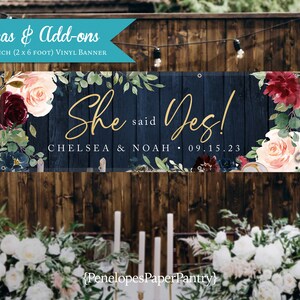 May include: A navy blue and white floral wedding banner with the text "She said Yes!" in gold script. The banner also includes the names "Chelsea & Noah" and the date "09.15.23".