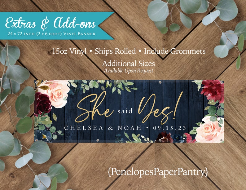 May include: A navy blue vinyl banner with a rustic woodgrain background and floral accents. The banner says "She said Yes!" with the names "Chelsea & Noah" and the date "09.15.23". The banner is 24 inches by 72 inches.