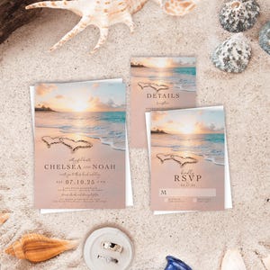 May include: A wedding invitation suite featuring a beach sunset scene with two hearts drawn in the sand. The cards include text for Chelsea and Noah, details, and an RSVP. A small white ring dish holds two silver rings, and a blue glass bottle is visible.
