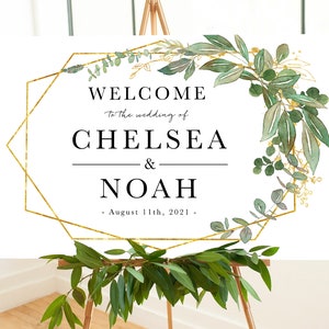 May include: A white welcome sign with a gold geometric frame and green foliage. The text reads "Welcome to the wedding of Chelsea & Noah - August 11th, 2021 -"
