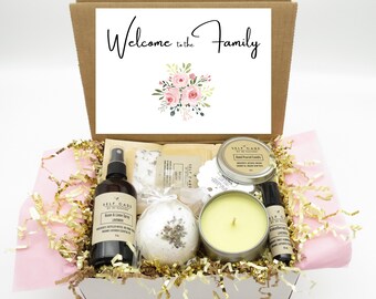 May include: A gift box containing a room and linen spray, a bath bomb, a hand-poured candle, and an aromatherapy roller. The box lid features the text Welcome to the Family with a pink floral illustration. The items rest on gold and pink paper filler.