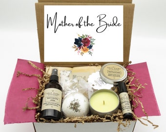 May include: A gift box labelled Mother of the Bride containing a room and linen spray, a bath bomb, a hand-poured candle in a metal tin, and an aromatherapy oil roller. The box is lined with magenta tissue paper and brown paper filler.