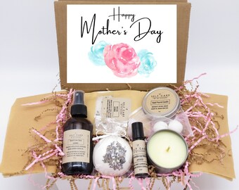 May include: A gift box filled with self-care items for Mother's Day. The box contains a body and love spray, a bath bomb, a hand-poured candle, a rollerball essential oil, and a bar of soap. The box is decorated with pink and brown shredded paper.